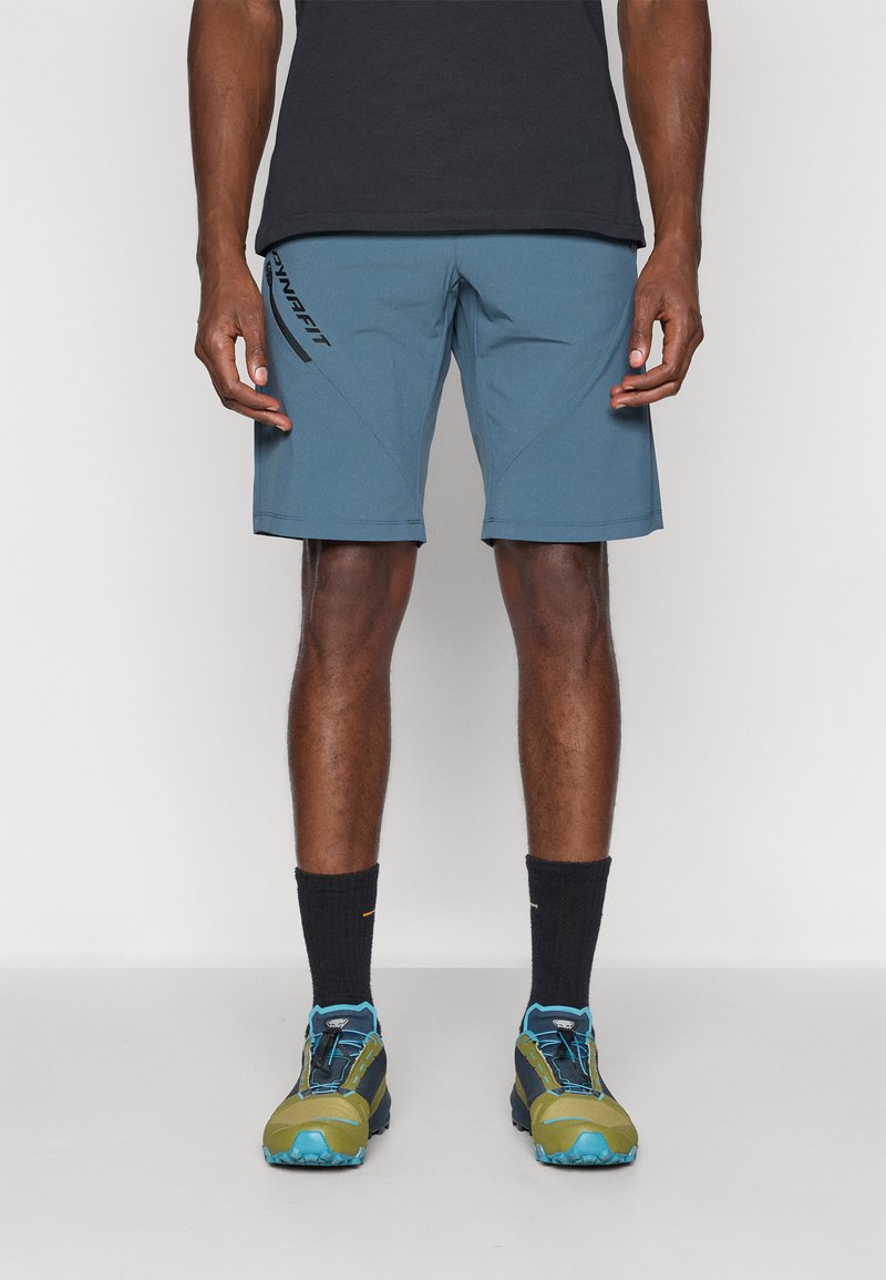 Μπλε σορτς πεζοπορίας με γωνιαστές ραφές και μαύρο λογότυπο στο μπροστινό μέρος. Φορεμένα με μαύρο πουκάμισο, μαύρες κάλτσες και πολύχρωπα παπούτσια πεζοπορίας.