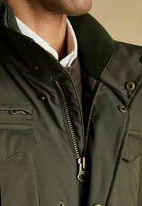 Ciemnozielona kurtka z wysokim kołnierzem, zapinana na zamek, z widocznymi szwami, wykończona mosiężnymi elementami oraz wieloma kieszeniami.