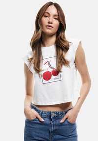 Camiseta corta blanca de manga corta, con un gráfico de cereza roja y texto en negro. Combinada con jeans de mezclilla azul de cintura alta.