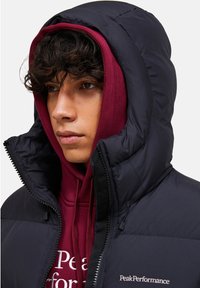 Marineblå pufferjakke med høj krave og hætte, lagdelt over en maroon hoodie med hvid tekst. Har lynlås og quiltet tekstur.