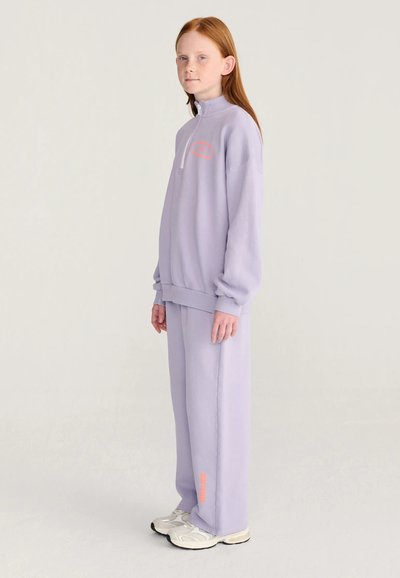 Enfant aux cheveux roux portant un survêtement lavender ample et des baskets blanches, debout de côté sur un fond clair uni.