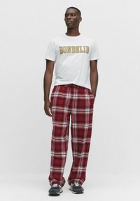 Vit T-shirt med texten "BONDED" i gult, kombinerad med röd- och vitrutiga pyjamasbyxor. Svarta sneakers syns vid fötterna.