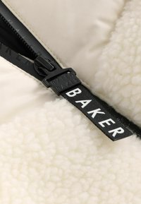 Kurtka puchowa w kremowym kolorze z puszystą teksturą, z czarnym zamkiem błyskawicznym i tkanym metkiem, na którym w białych literach widnieje napis "BAKER".