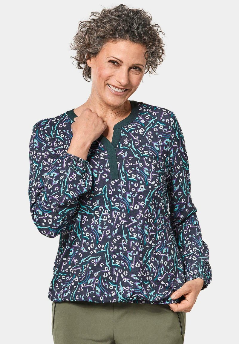 Langärmlige Bluse aus navyfarbenem Stoff mit floralem Muster in Weiß und Türkis. Verfügt über einen V-Ausschnitt mit dunkelgrünem Besatz und gerafften Ärmeln.