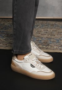 Sneakers bianchi e beige con pelle testurizzata, dettagli perforati e una robusta suola in gomma marrone chiaro. I lacci sono dello stesso beige con occhielli metallici.
