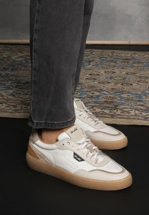 Sneakers bianchi e beige con pelle testurizzata, dettagli perforati e una robusta suola in gomma marrone chiaro. I lacci sono dello stesso beige con occhielli metallici.
