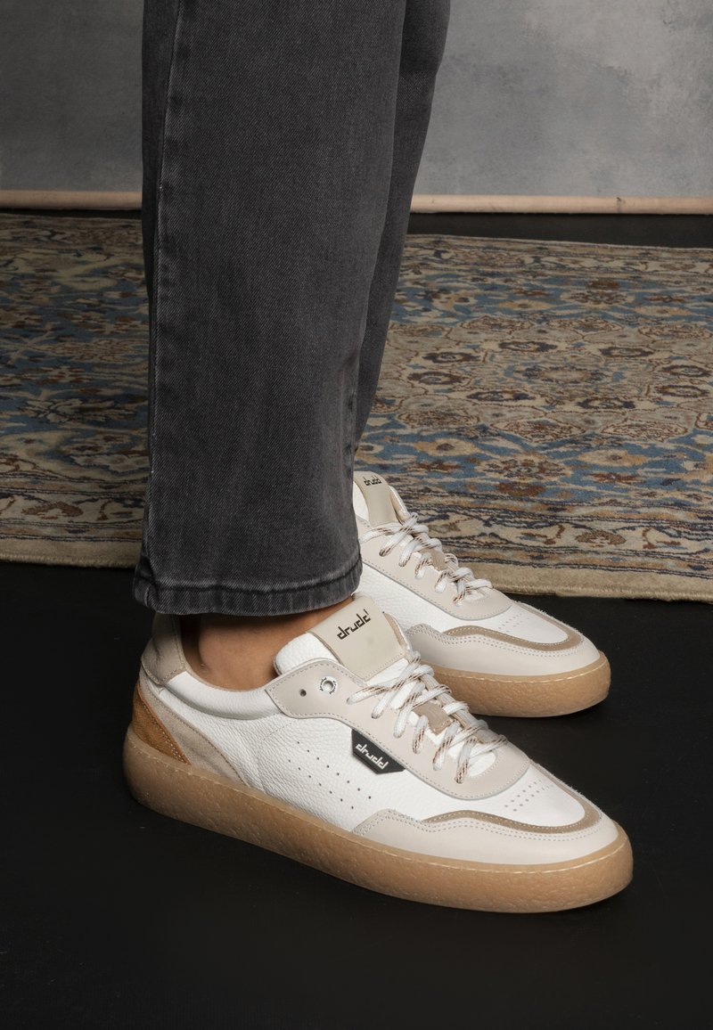 Sneakers bianchi e beige con pelle testurizzata, dettagli perforati e una robusta suola in gomma marrone chiaro. I lacci sono dello stesso beige con occhielli metallici.