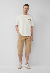 Beige Cargo-Shorts mit Taschen, kombiniert mit einem weißen Baumwoll-T-Shirt mit einem bunten Blumenmuster. Cremefarbene und braune Sneaker runden den Look ab.