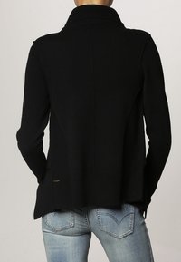 Svart stickad tröja med hög krage, ribbad textur och sidosprund. Bakifrån visar den rena linjer och en subtil logotypdetalj.