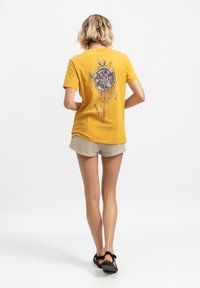 Maglietta di cotone gialla con una grafica circolare sul retro, che presenta dettagli di orologio e frecce. Abbinata a pantaloni corti beige e sandali neri.