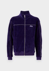 Lilla velour zip-up jakke med høy krage, sølvfargede aksentstriper og brodert logo. Har to frontlommer og ribbede mansjetter.
