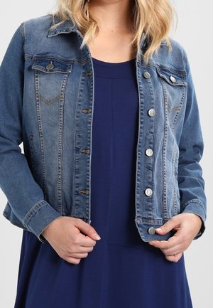 Frau trägt eine blaue Jeansjacke über einem dunkelblauen Kleid und hält den Saum der Jacke mit beiden Händen.