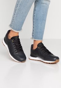 Svarta Nike sneakers har en slät läderöverdel, en vit mellanläggssula och en gummisula, med svarta skosnören och minimal märkning på tungan.