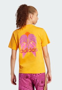 Žluté bavlněné tričko s potiskem palmy a tukana, s růžovými akcenty a tučným nápisem "adidas" na zadní straně.