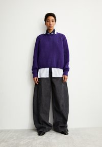 Un pull en tricot épais violet porté par-dessus une chemise blanche à col, associé à un pantalon en denim noir à jambes larges. Des chaussures noires avec des clous argentés.