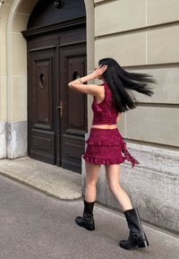 Burgundy lace crop top with ruffled accents and matching mini skirt, paired with black knee-high boots, walking past an ornate wooden door.