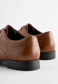 Brown leather shoes with a smooth finish, featuring a textured panel, contrasting stitching, and a black rubber sole. Close-up of heel details.