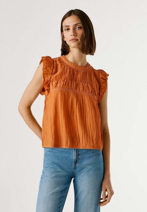 Jeune femme portant un haut sans manches rouille à volants avec des plis, associée à un jean bleu clair, debout devant un fond uni.