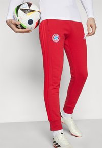 Røde joggebukser med tre vertikale striper nedover hver side, med Bayern München-logoet på venstre lår. Teksturen er glatt bomull.