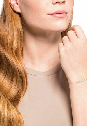 Pulsera de oro con tres cuentas azul claro, llevada en una mano; el modelo tiene el cabello rojo ondulado y viste una blusa beige. Textura suave y diseño delicado.