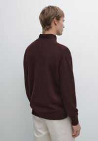 Bordowy sweter z dzianiny z wysokim kołnierzem, żebrowanym dołem i bezszwowymi ramionami, noszony na jasnobeżowych spodniach. Miękka tekstura, casualowy krój.