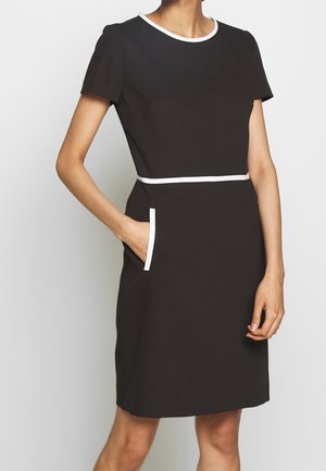 Frau trägt ein kurzärmliges schwarzes Kleid mit weißem Besatz am Ausschnitt, an der Taille und an der Tasche, hält eine Hand in der Tasche.