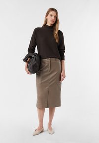 Frau in einem dunkelbraunen Pullover und einem taubenfarbigen Leder-Midirock, hält eine große geflochtene Clutch und steht vor einem schlichten hellen Hintergrund.