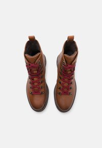 Botas de senderismo de cuero marrón con superficie texturada, cordones rojos, ojales de metal y tira de agarre en el talón. Vista superior que muestra una disposición simétrica.