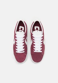 Burgundy suede sneakers with white laces and accents, featuring perforated toe area and a logo on the tongue. Classic low-top design.