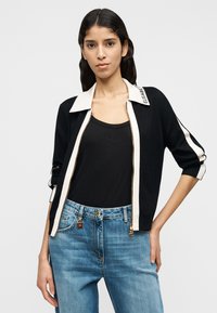 Μαύρη ζακέτα με λευκό γιακά, κλείσιμο με φερμουάρ και ρίγες στο πλάι. Φοριέται πάνω από μαύρο τοπ. Συνδυάζεται με ανοιχτόχρωμο μπλε τζιν με χρυσές λεπτομέρειες.