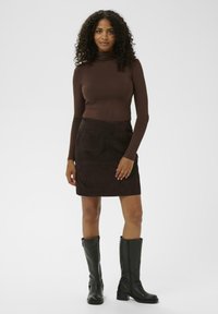 Brun langærmet turtleneck-top partnered med en mørkebrun skirt i ruskind, der har forlommer; sorte knæhøje støvler med en kraftig hæl.