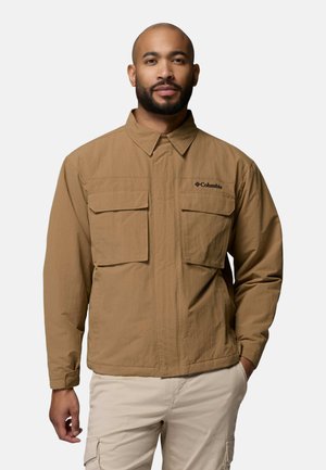 Mann trägt eine beige Columbia-Jacke mit großen Brusttaschen und hellbeigen Hosen, steht mit einer Hand in der Tasche vor einem schlichten Hintergrund.