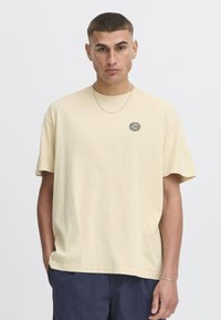 Kortærmet, lys beige bomulds-t-shirt med rund halsudskæring og et lille, sort solgrafik på venstre bryst. Glat tekstur.