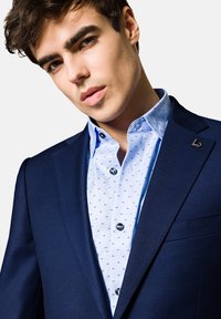 Granatowa marynarka wykonana z usztywnionego materiału, pod którą znajduje się jasnoniebieska koszula w wzory, akcentowana metalowymi guzikami i przypinką z logo.