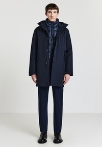 Cappotto impermeabile blu navy con chiusura a bottoni, grande cappuccio e tasche laterali, indossato sopra un giubbotto imbottito. Abbinato a pantaloni scuri e stivali coordinati.