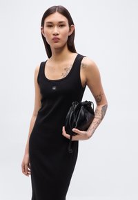 Μαύρο αμάνικο φόρεμα με εφαρμοστή σιλουέτα, με τετράγωνο ντεκολτέ και λογότυπο. Στο χέρι κρατιέται μια μικρή μαύρη δερμάτινη τσάντα με κορδόνι.