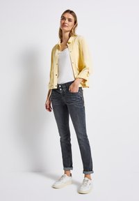 Ljusgul knappad jacka över en vit skjorta, ihop med mörkgrå jeans och vita sneakers. Jeansen har sidoknappar och uppvikta ärmar.