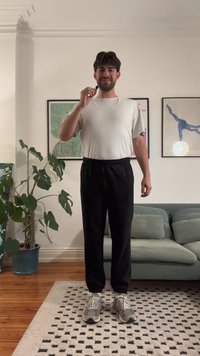 Person wearing a white t-shirt and black elastic-waist trousers, standing on a patterned rug, with gray sneakers, and indoor plants in background.
