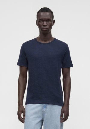 Junger Mann mit dunkler Haut, der ein marineblaues kurzärmeliges T-Shirt und hellblaue Jeans trägt, steht vor einem schlichten weißen Hintergrund.