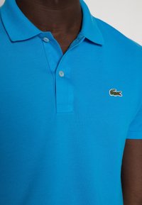 Polo bleu en coton texturé, doté d'une patte de boutonnage à trois boutons et d'un petit logo crocodile brodé sur le côté gauche de la poitrine.