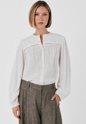 Femme aux cheveux blonds courts portant un chemisier blanc boutonné et un pantalon gris taille haute, une main dans la poche.