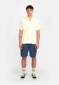 Kurzärmliges, gelbes Hemd mit Knöpfen und einer Tasche, kombiniert mit navyblauen Shorts. Die Socken haben mehrfarbige Streifen und schwarze Sneakers.