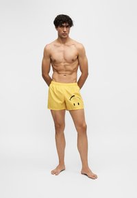 Jeune homme aux cheveux bouclés portant un short de bain jaune avec un visage souriant noir, debout pieds nus sur un fond blanc uni.