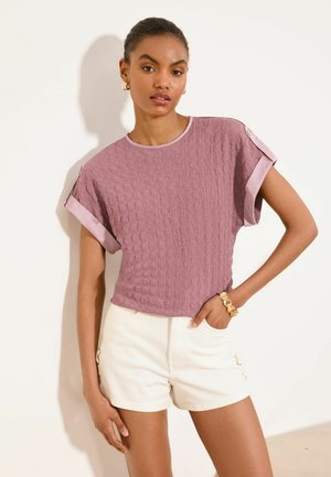 Nő texturált mályva színű rövid ujjú felsőt visel mandzsetta részletekkel és magas derekú fehér rövidnadrágot, és az egyik kezét zsebébe teszi pózolás közben.