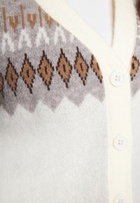 Cárdigan en crema con un patrón de punto texturizado que presenta formas de diamante en marrón y gris; cinco botones blancos; material suave y cálido.