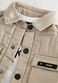 Beige quiltet jakke med sorte trykknapper, en brystlomme med et "Baker by Ted Baker" mærkat, over et hvidt plagg med sort tekst.