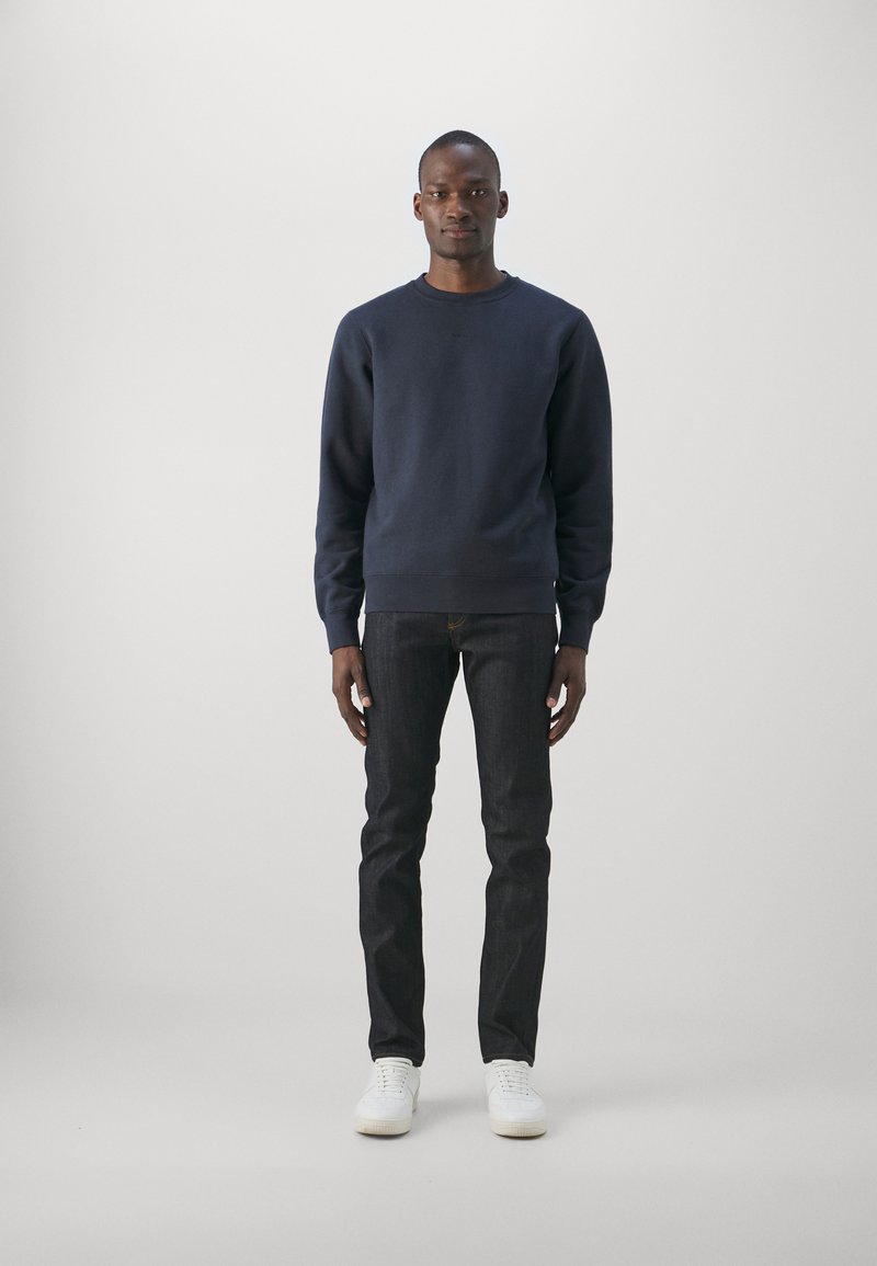 Marineblaues Sweatshirt aus weichem Material, mit Rundhalsausschnitt, gerippten Bündchen und Saum. Kombiniert mit dunklen Jeans und weißen Sneakers, vor einem hellen Hintergrund stehend.