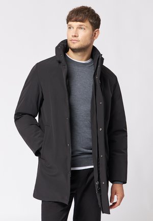 Sort isoleret frakke med lynlås foran, høj krave og trykknapdetaljer. Viser en glat tekstur og let tapered pasform.