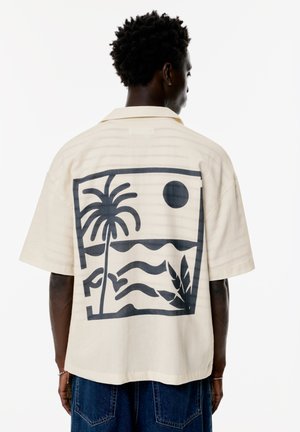 Short-sleeve, cream shirt with a black palm tree and sunset graphic on the back. Features a relaxed fit and soft, textured fabric.