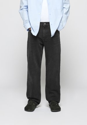Black denim jeans with a loose fit, featuring a button and zip fly, worn with a light blue button-up shirt and black slip-on shoes.